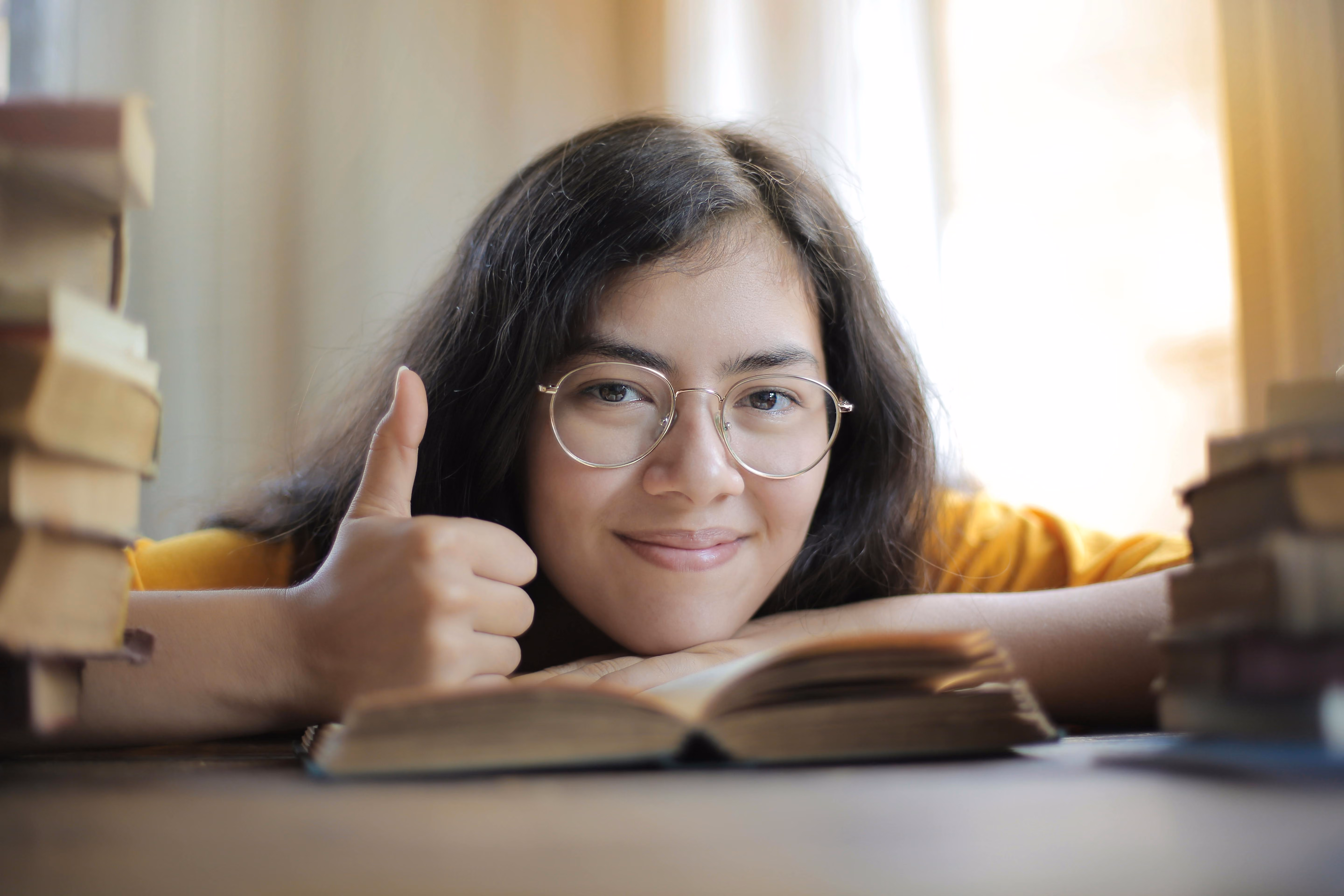 Student giving thumbs up while studying among stacks of books