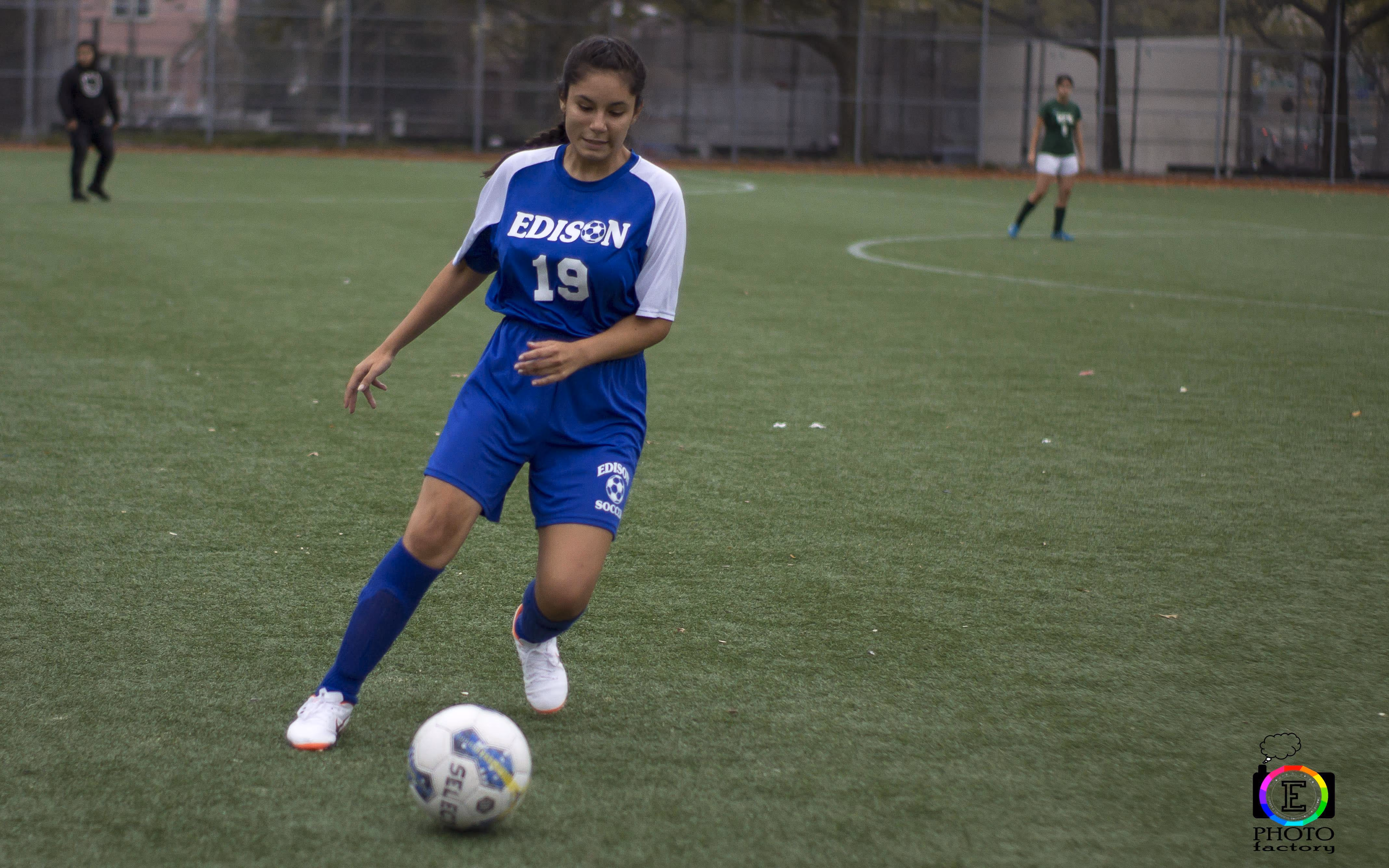 Female soccer player in blue Edison uniform with number 19 dribbling a soccer ball on a green field.
