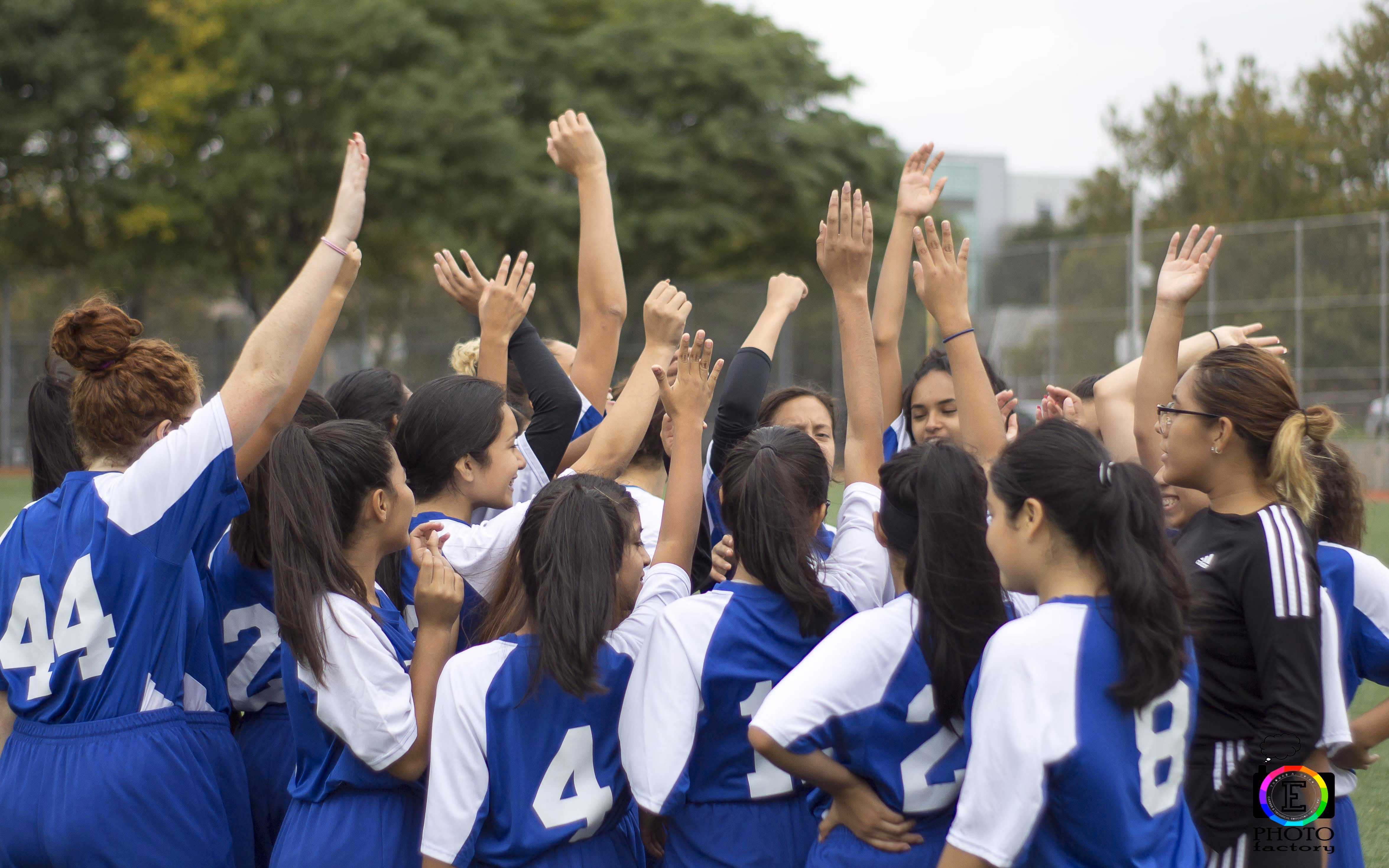 Girls soccer team wearing blue and white uniforms raising their hands in a group huddle on a field.