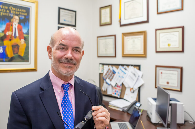 Smiling middle-aged man in a suit and blue polka dot tie holding glasses in an office with framed certificates and a colorful painting in the background.