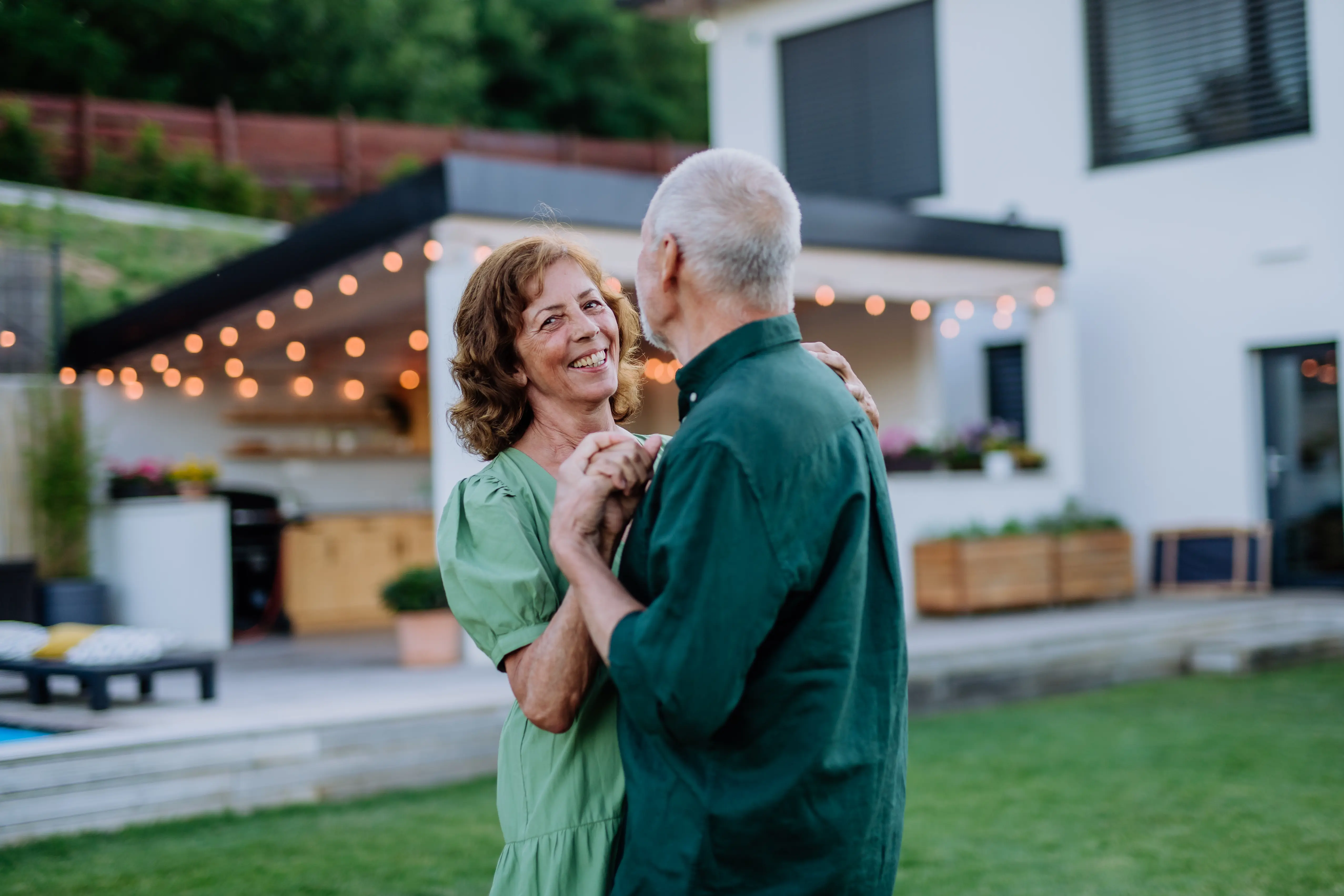 Smiling elderly couple dancing together outdoors in a backyard with string lights and modern house in the background.