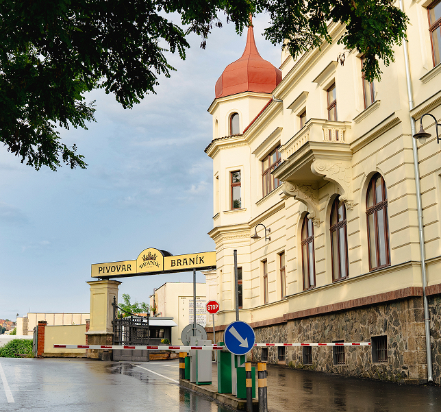 Vstupní brána pivovaru Braník vedle oranžově zastřešené historické budovy za mokré silnice s dopravními značkami.