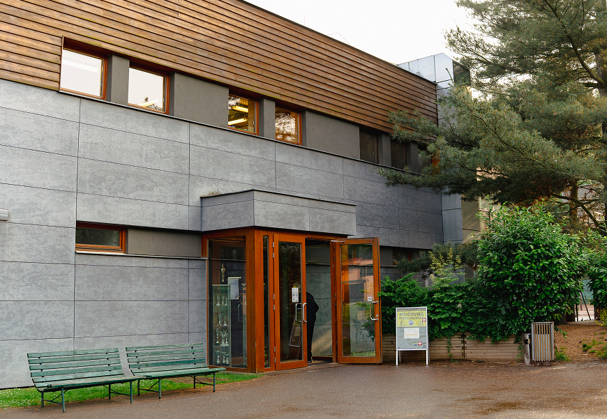 Moderní budova s šedými stěnami, dřevěným obkladem nahoře, skleněnými dveřmi a zelenými lavičkami venku před vchodem, obklopená stromy a keři.