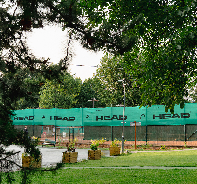 Venku tenisové kurty s modrozelenými bannery HEAD obklopené zelení a stromy.