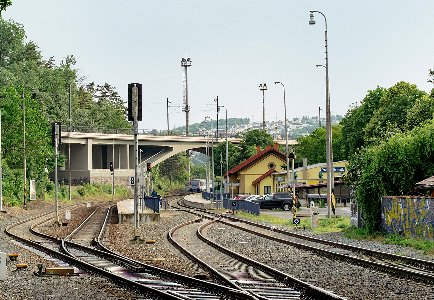 Železniční koleje vedoucí k malému nádraží obklopenému zelení a auty, s mostem v pozadí.