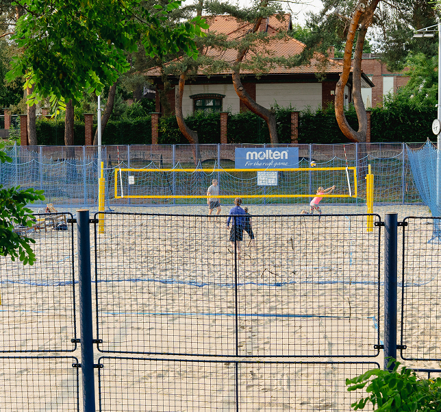 Lidé hrající plážový volejbal na pískovém hřišti s modrou sítí a žlutým volejbalovým základem, v pozadí stromy a dům.
