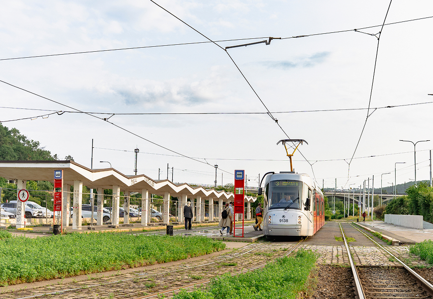 Tramvaj přijíždějící na zastávku s několika čekajícími lidmi a zeleným porostem podél kolejí.