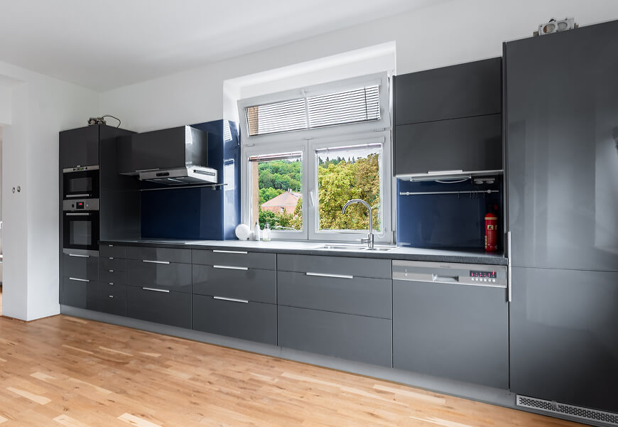 Moderní kuchyňská linka s lesklými tmavě šedými skříňkami, nerezovým dřezem pod oknem a světlou dřevěnou podlahou.