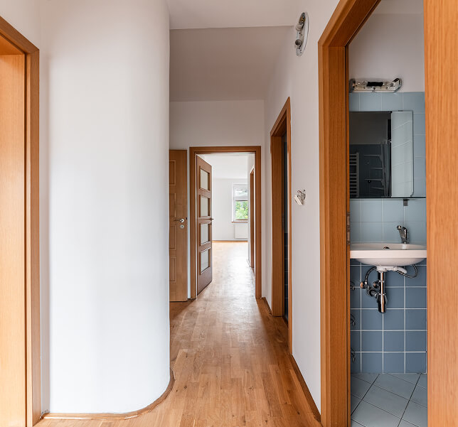 Chodba s dřevěnou podlahou a bílými stěnami vede ke dveřím místnosti se stejně dřevěnými dveřmi, vpravo je koupelna s modrými dlaždicemi a umyvadlem.