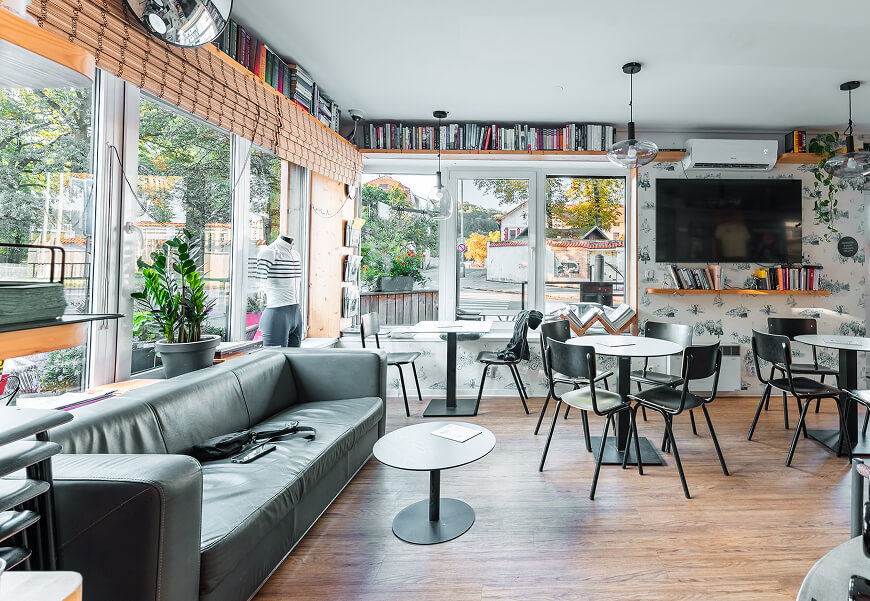 Moderní kavárna s koženou pohovkou, malými kulatými stolky, knihovnou podél horní části stěn a velkými okny s výhledem na venkovní zeleň.