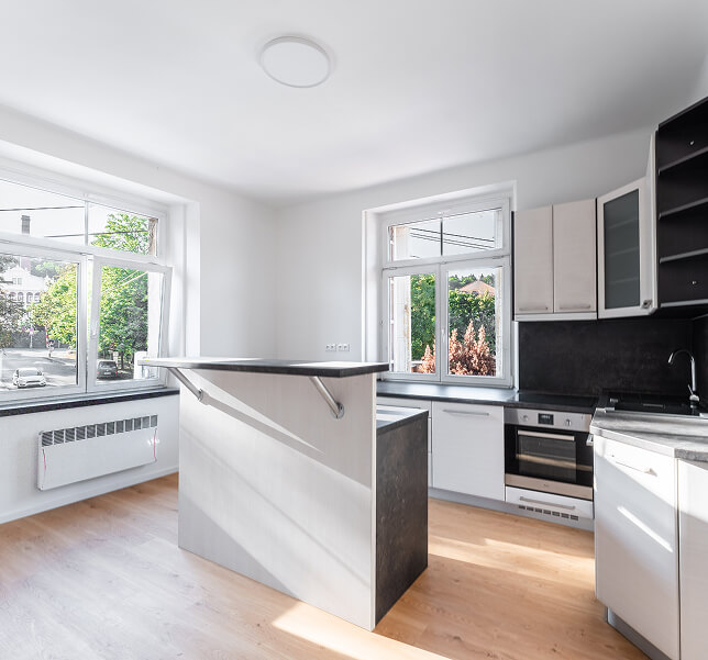 Světla moderní kuchyň se světlými skříňkami, černou pracovní deskou a dvěma okny s výhledem na zelené stromy.