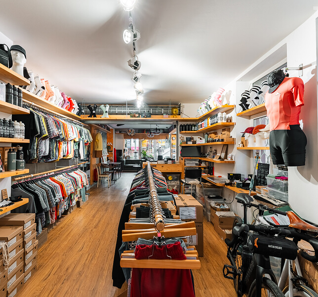 Interiér moderního obchodu s cyklistickým vybavením a oblečením, řada oblečení na ramínkách a sportovní kolo vpravo.