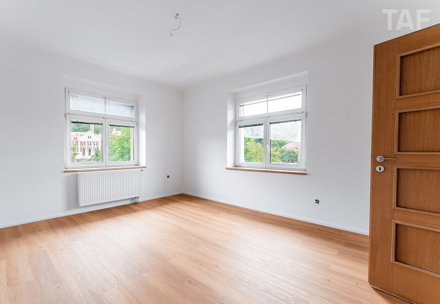 Prázdný světlý pokoj s dřevěnou podlahou, dvěma okny s výhledem na zeleň a částečně otevřenými žaluziemi, vedle nich je otevřené dřevěné dveře.