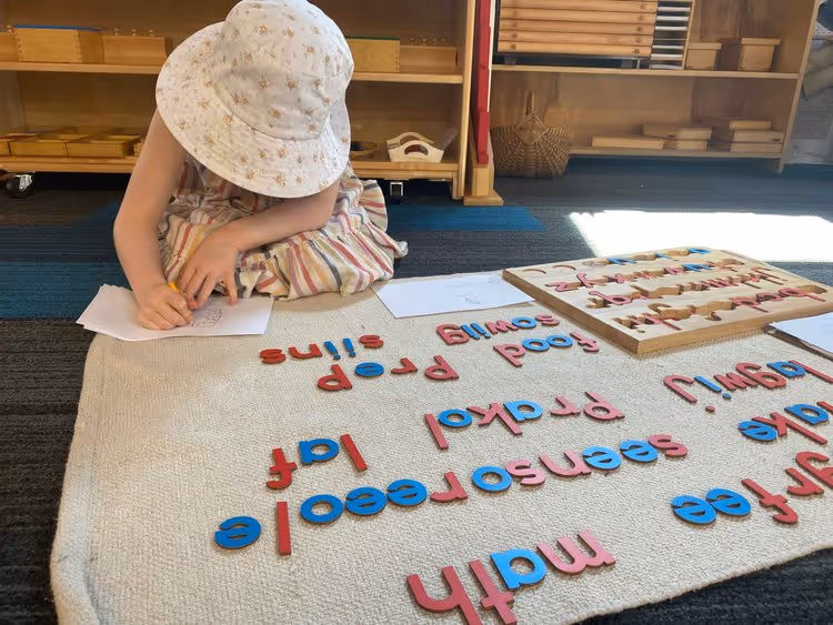 Child wearing a floral bucket hat and striped dress sitting on a carpet and writing on paper, with colorful wooden letter blocks scattered on the carpet.
