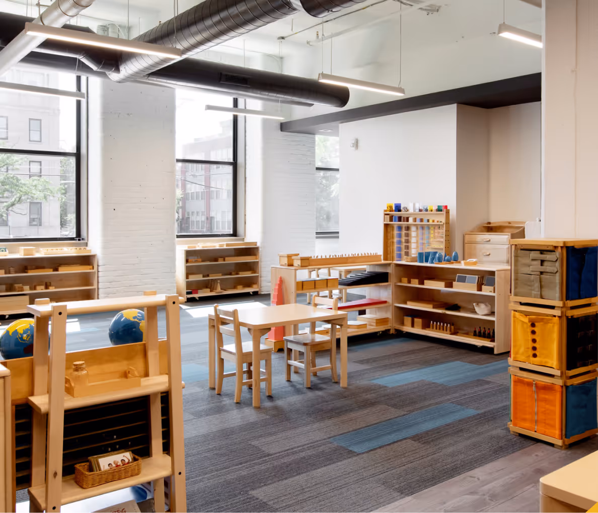 Bright classroom with wooden shelves, tables, and chairs, natural light from large windows, and educational materials.