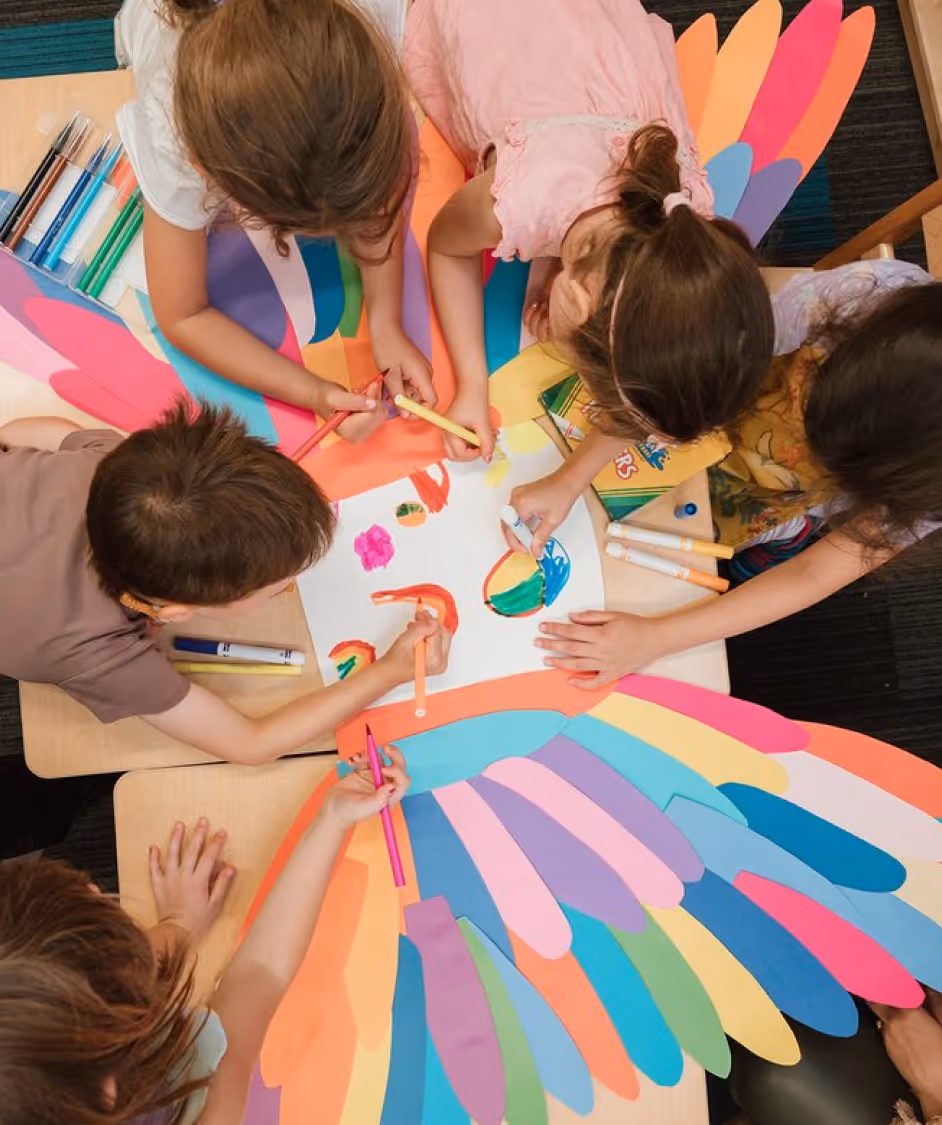 Children gathered around a table drawing and coloring on a large paper with colorful paper wings underneath.