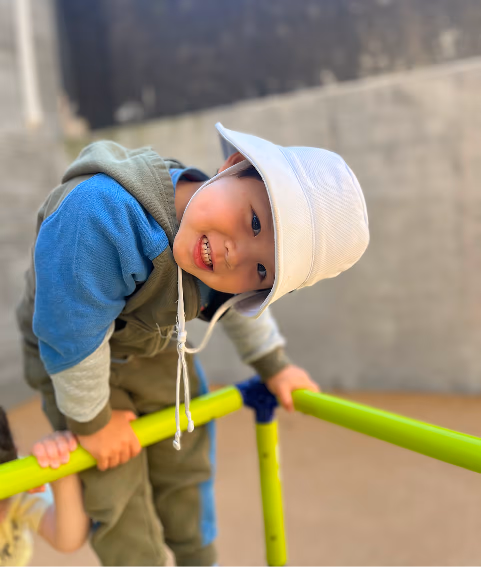 Smiling young child wearing a white hat and blue and green outfit climbing on bright green playground bars.