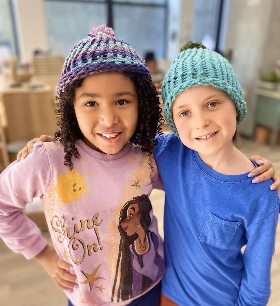 Two children wearing knitted hats smiling with their arms around each other indoors.