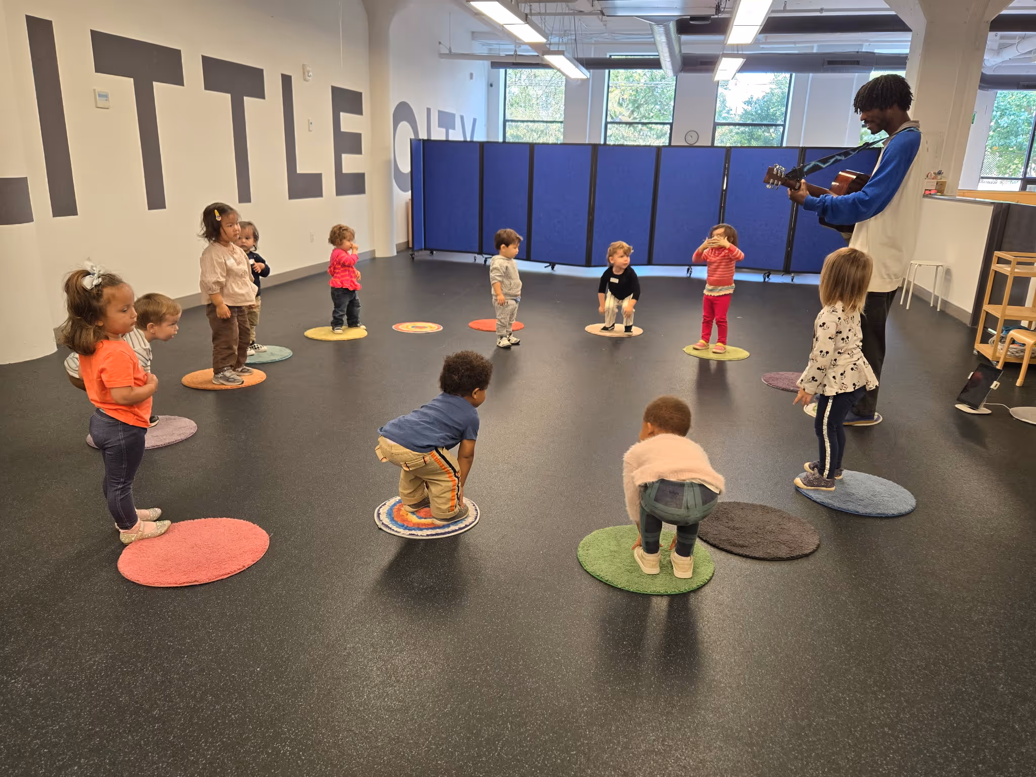 Group of young children standing or crouching on colorful circular mats in a classroom while an adult plays guitar.
