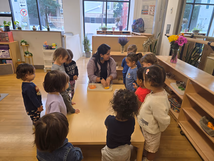 Teacher seated at a table demonstrating an activity to a group of young children standing around her in a bright classroom.