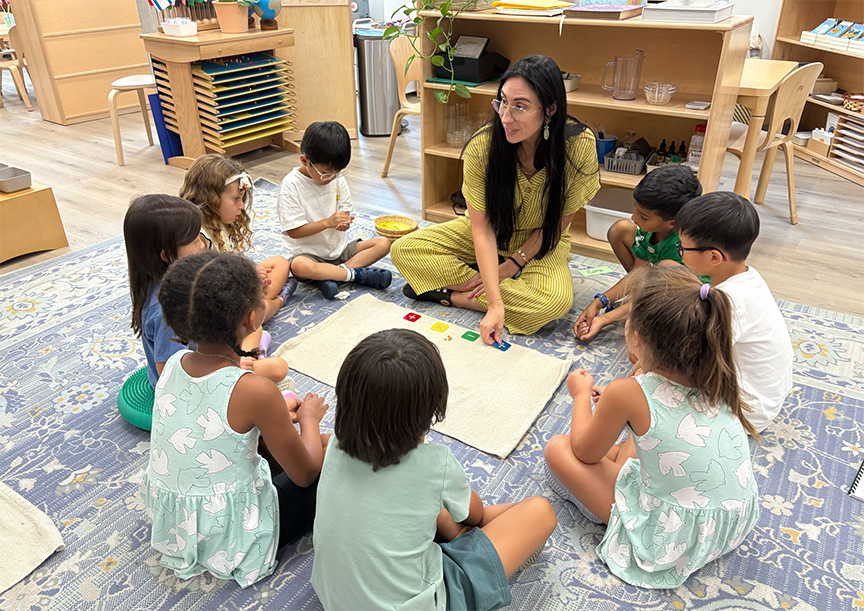 Teacher sitting on the floor with a group of young children in a circle, engaging in an educational activity using colored cards on a beige mat.