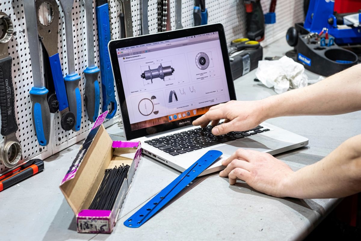 Eine Person arbeitet in einer Fahrradwerkstatt an einem aufgeklappten MacBook Pro, auf dessen Bildschirm technische Zeichnungen von Fahrradteilen zu sehen sind. Auf der Werkbank liegen Werkzeuge, ein blauer Metallstreifen und eine geöffnete Schachtel mit Kabelbindern; im Hintergrund hängen weitere Werkzeuge an einer Lochwand.