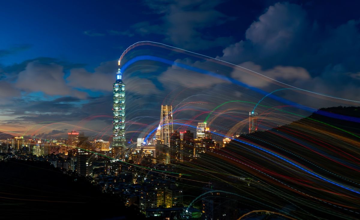 Nachtansicht einer beleuchteten Großstadt mit einem hohen, hell erleuchteten Wolkenkratzer im Zentrum. Farbige Lichtspuren in Bögen ziehen über den Himmel und durch die Stadt und symbolisieren digitale Vernetzung, Datenströme oder internationale Kommunikation.