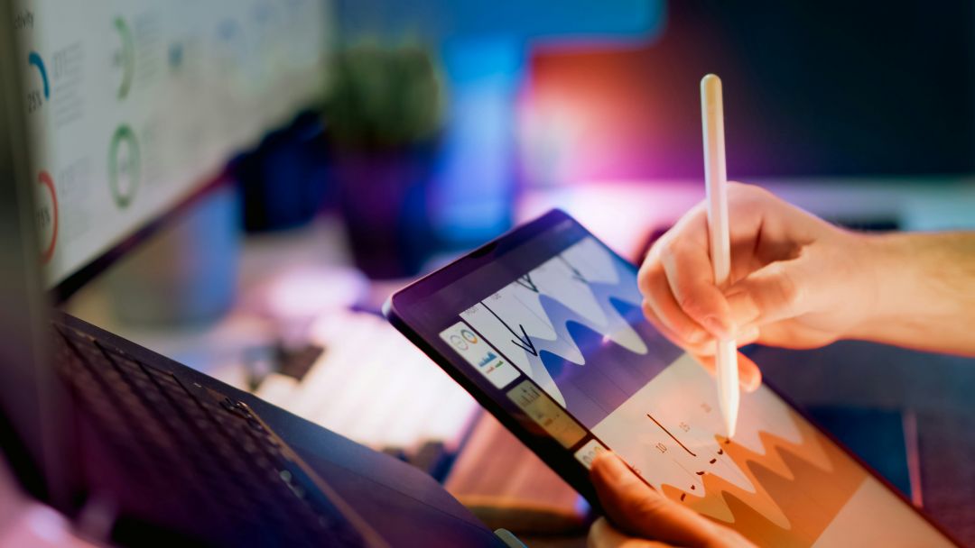 Hand bedient Tablet mit Diagrammen und Stift neben Laptop.