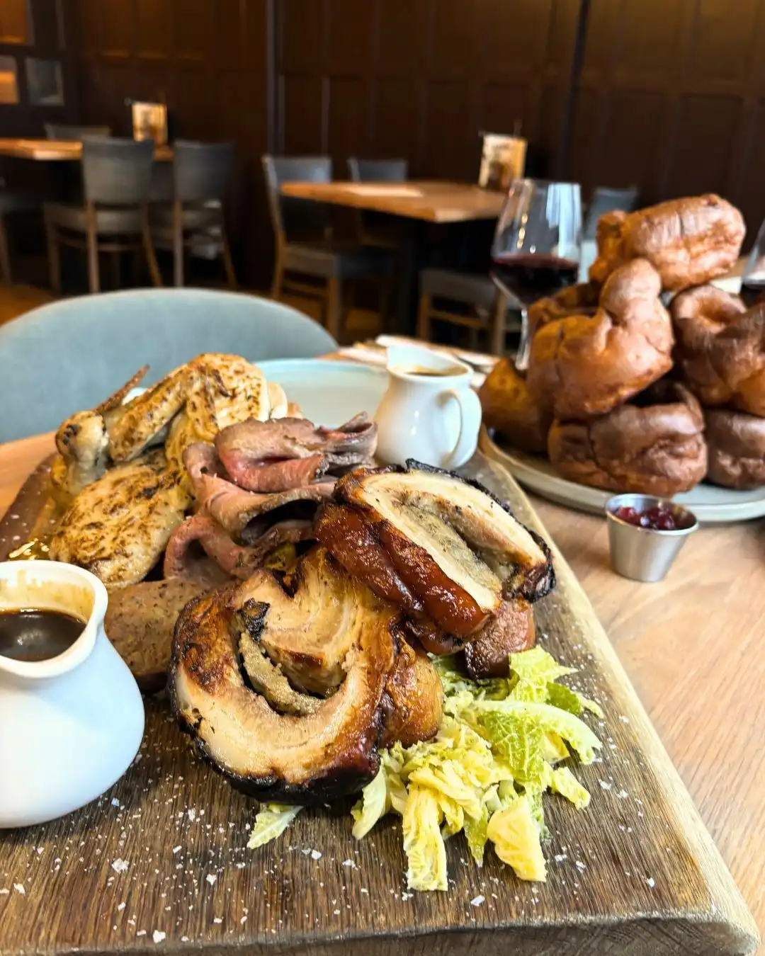 Roast dinner on a wooden board with various meats, gravy, and cabbage. Yorkshire puddings and a glass of red wine are in the background on a table.