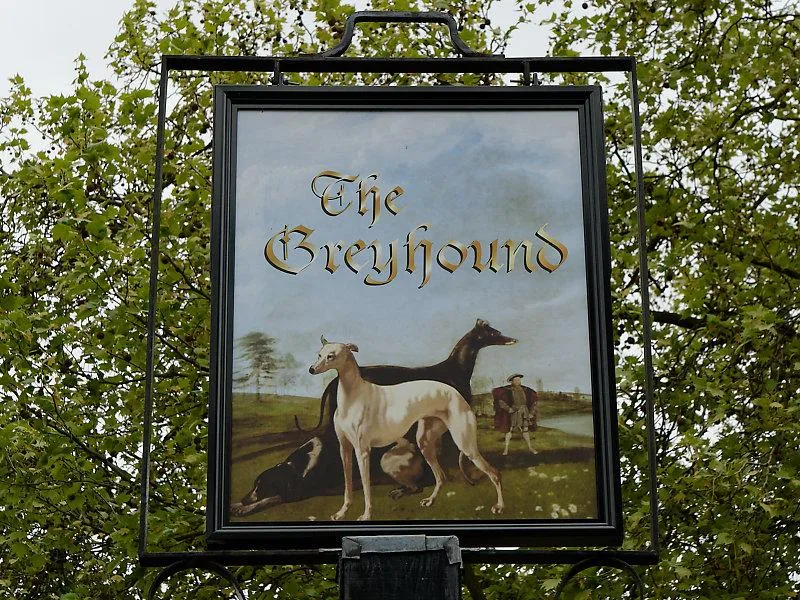 Sign for "The Greyhound" pub featuring a painted scene of greyhounds in a lush field, with muted earth tones and a calm, pastoral atmosphere.