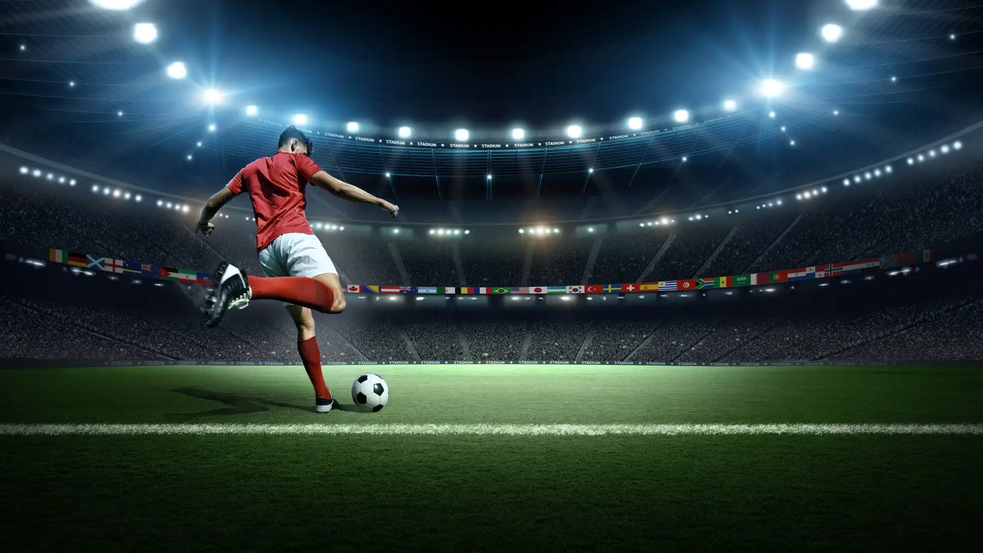 Soccer player in red shirt and white shorts kicking ball on a floodlit stadium field.