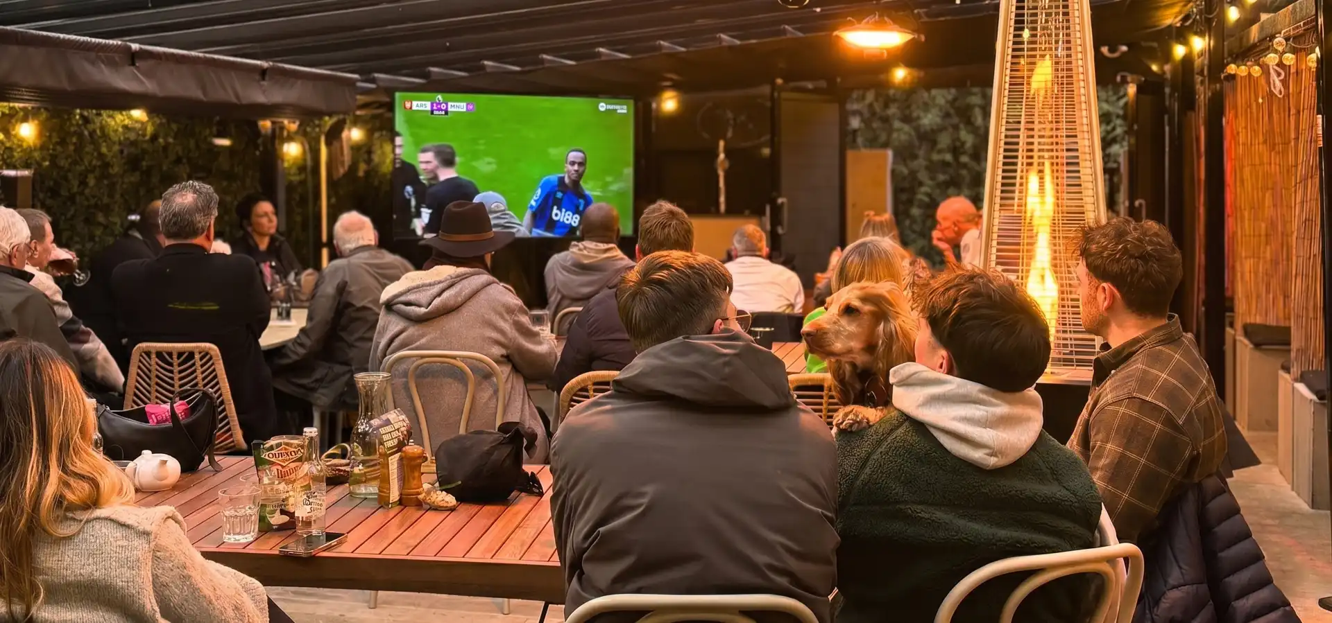 People sit at tables in an outdoor bar or restaurant watching a soccer match on a large screen; some are eating, drinking, and one person has a dog on their lap.