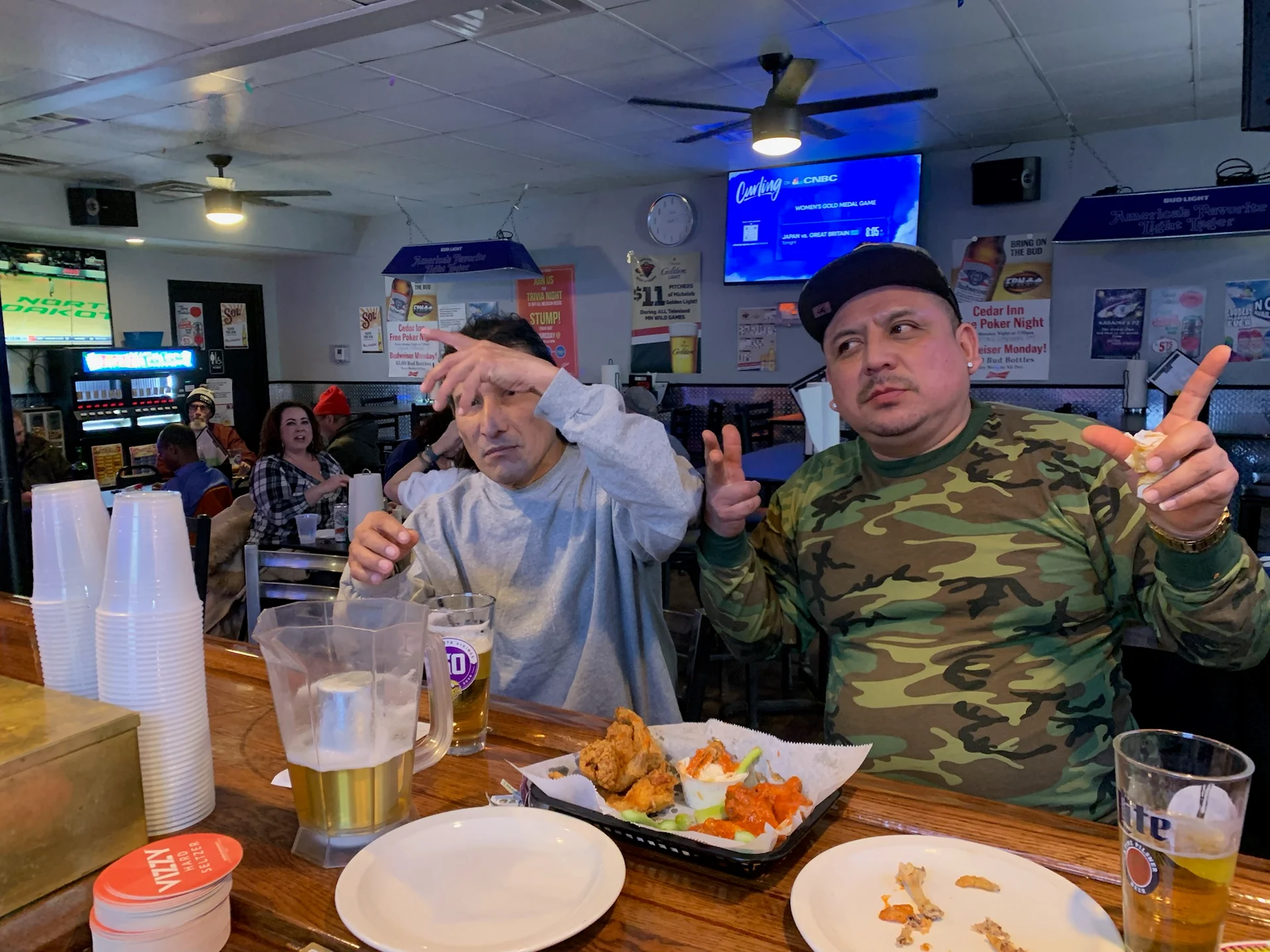 Two men sitting at a bar with a plate of food and drinks.