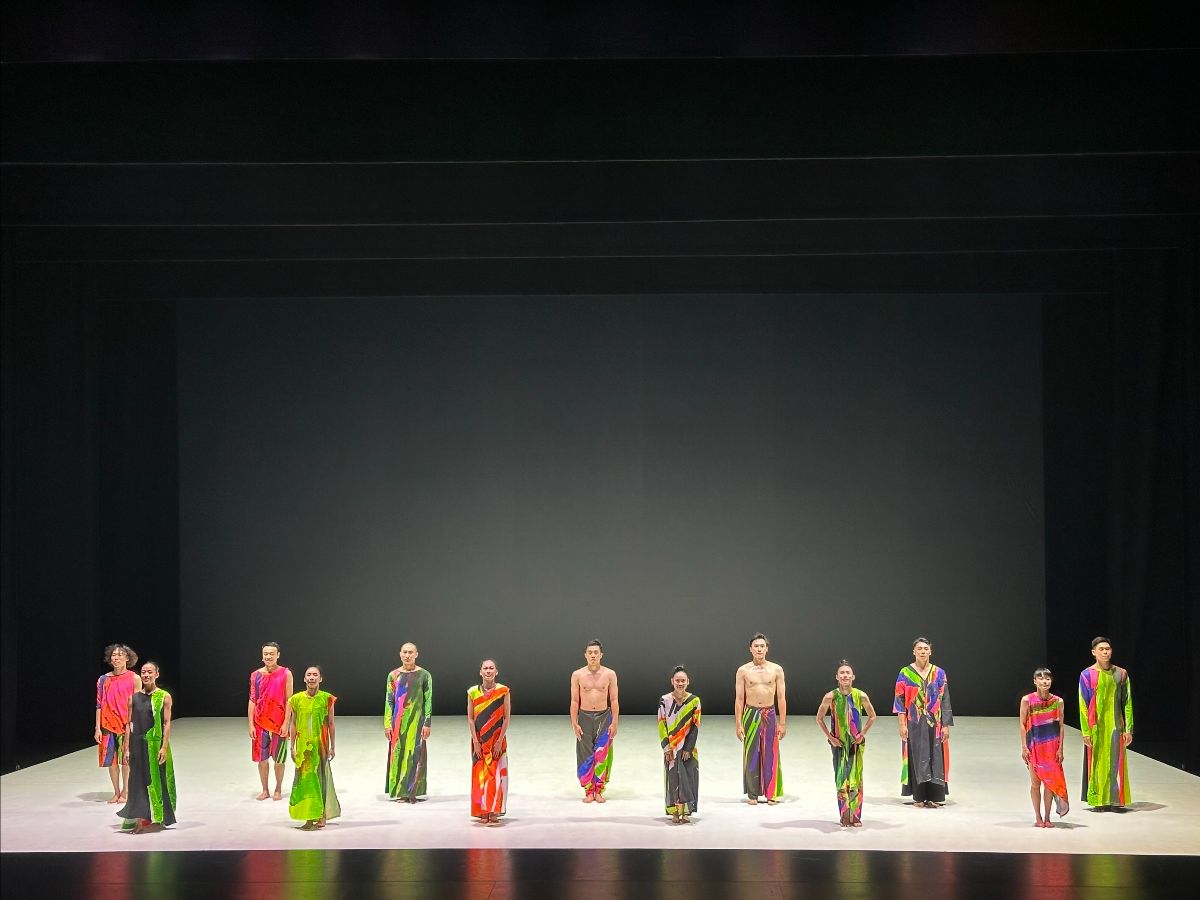 13 members of Cloud Gate Dance Theatre of Taiwan wear fluorescent-colored outfits and stand in two rows on a white stage floor with a black backdrop at the conclusion of a performance of 13 TONGUES in Ann Arbor, Michigan.