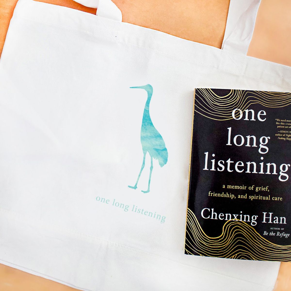 White tote bag on teal-colored crane and the words "one long listening." A copy of the book “one long listening” by Chenxing Han rests on the bag. The book cover is black with wavy gold threads framing the title and author name.