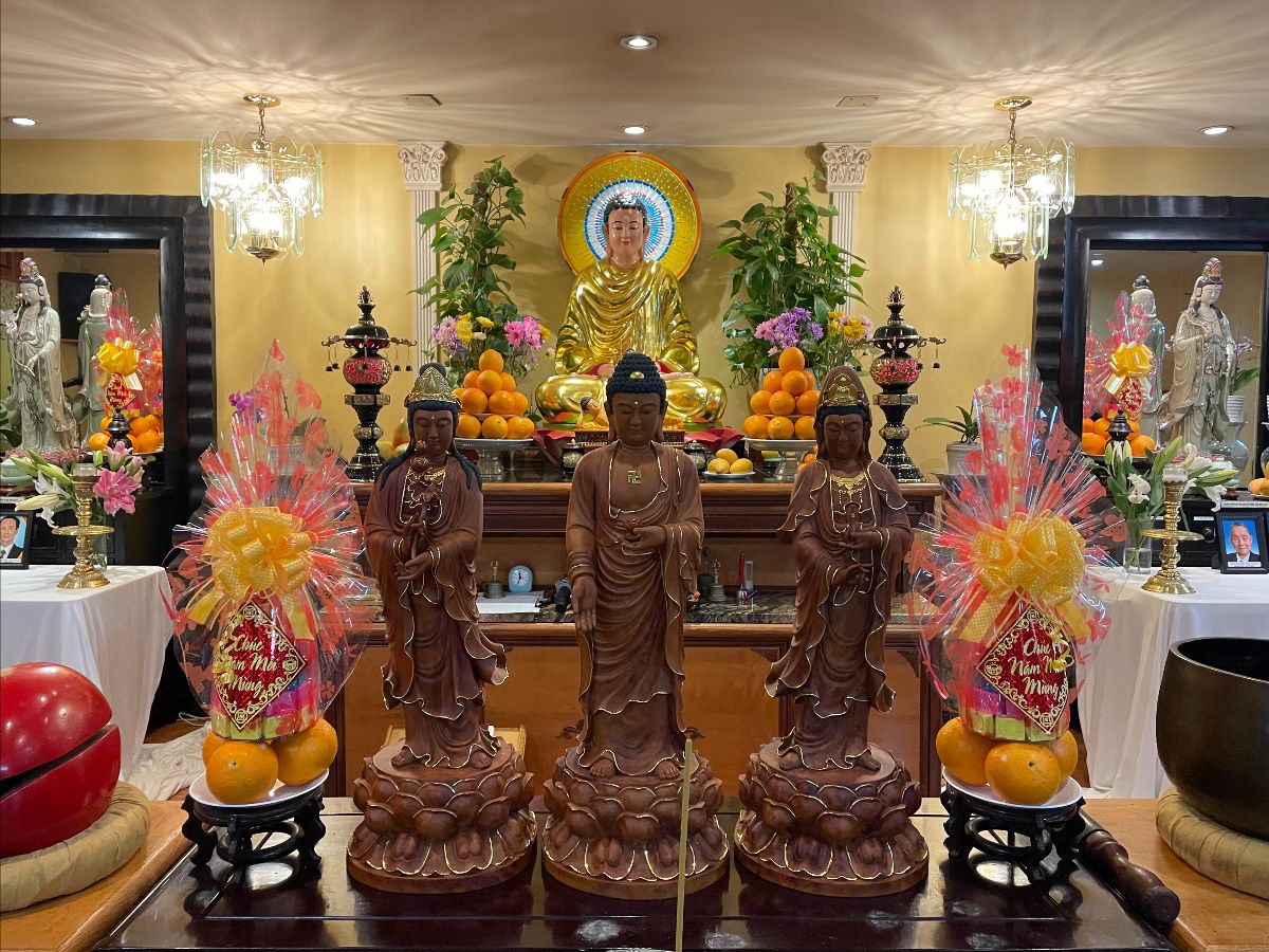 Multiple altars at a Vietnamese Buddhist temple in Lowell, Massachusetts. The altar in the foreground has three dark brown standing buddhas/bodhisattvas flanked by a pair of fruit offerings (decoratively wrapped oranges with the words “Chúc mừng năm mới”). There is red wooden fish (mõ) on the left and a bell and striker on the right. A larger altar in the back has a seated gold Buddha with a colorful halo behind his head. The seated Buddha is surrounded by multiple fruit offerings and flanked by a pair of incense burners. Two additional side altars in the back each have a standing white Quan Âm statue and a large framed mirror behind it. The wall is painted yellow and two brightly lit glass chandeliers hang from the ceiling.