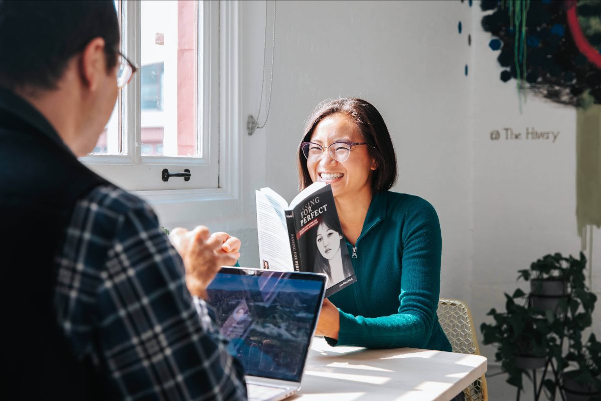 Chenxing Han and Trent Walker across from each other at a white desk by a window. Chenxing wears a turquoise zip-up sweater and is smiling and holding a copy of the book “Passing for Perfect.” The book depicts a young Asian woman with black hair on the cover. Trent is wearing a black vest over a plaid shirt and has an open laptop in front of him.