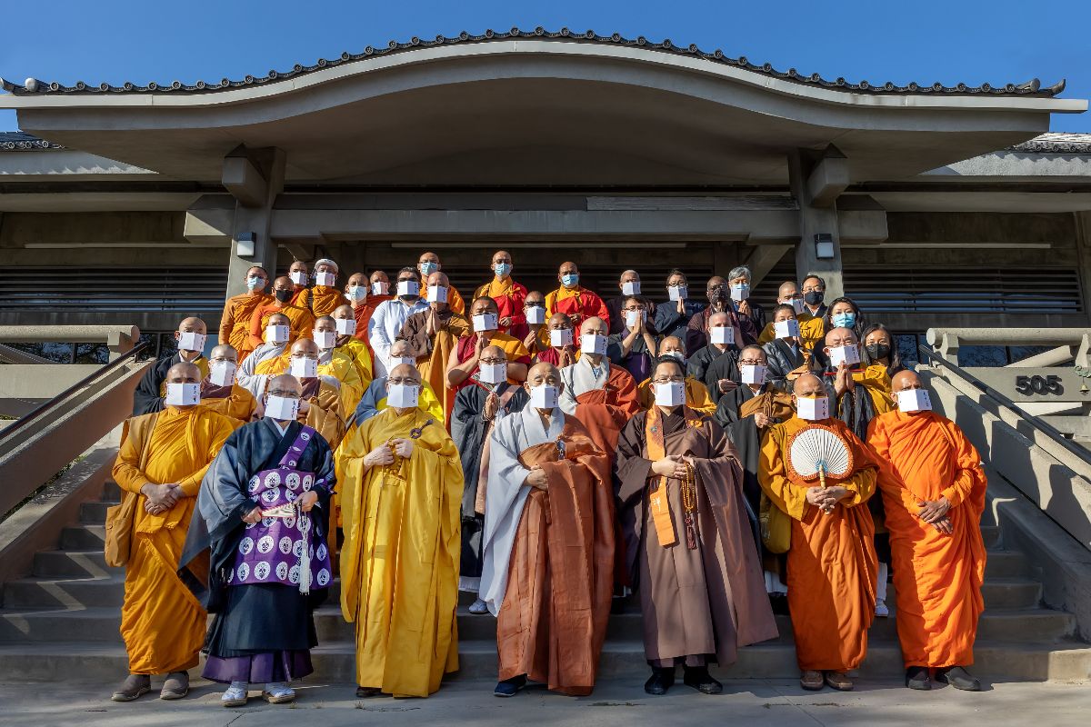 A group of forty-four Buddhist monastics and laypeople, most of whom are wearing religious robes, stand on the steps at the front entrance of Higashi Honganji Buddhist Temple in Los Angeles, California. Everyone is masked. Most attendees wear a white paper mask decorated on one side with vertical calligraphy in black ink.
