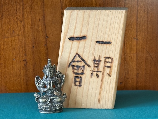 A small silver-colored statue of a multiarmed Buddha figure (Tara) sits on a teal-colored fabric book cover next to a block of balsa wood that has the characters 一期一會 (ichigo ichie) burned into it. The background is cherry-colored wood.