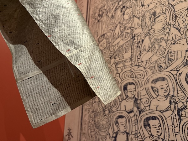 Sleeve of silver silk garment covered in tiny black Chinese characters against a background of multiple haloed Buddhist figures, photographed at the Princeton University Library exhibit Forms & Function: The Splendors of Global Book Making