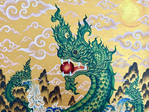 Closeup of a Thai painting depicting three scaly green nagas (dragon-like creatures) with flame-like ornamentation on their serpentine heads. The large naga in the foreground holds a red ball in its open mouth between its front teeth. The smaller nagas in the background on the left and right spew water from their mouths. The background shows brown mountains and white clouds against a golden sky, with a golden sun in the top right corner.