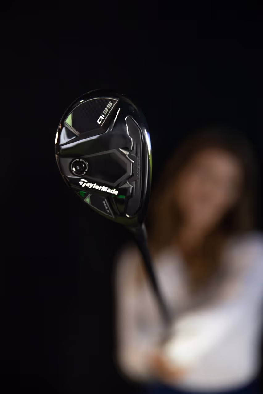 Blurred woman golfer in the background, with focus on the bottom of the TaylorMade Qi35 Rescue Hybrid.