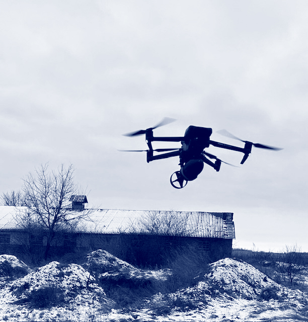 Drone hovering near snow-covered barn and rocky terrain on cloudy day