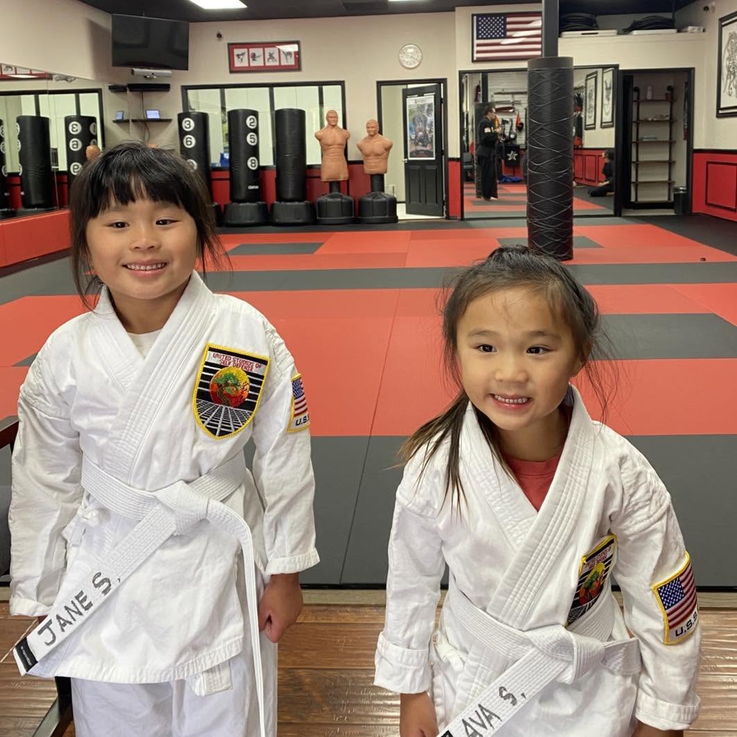 Two white belt kids karate students standing and smiling together at the entrance of USSD Irvine Woodbridge.