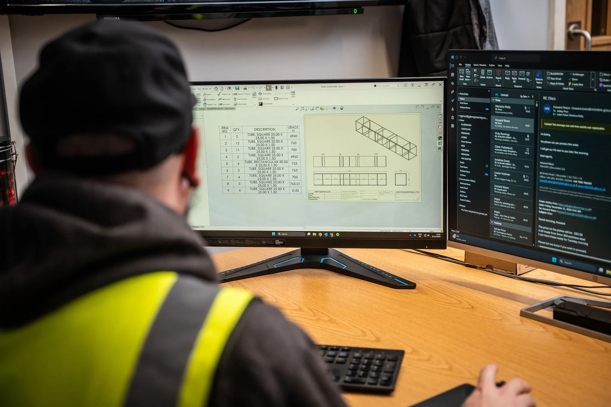 Man wearing a hat and safety vest working on CAD design and email on dual monitors at a desk.