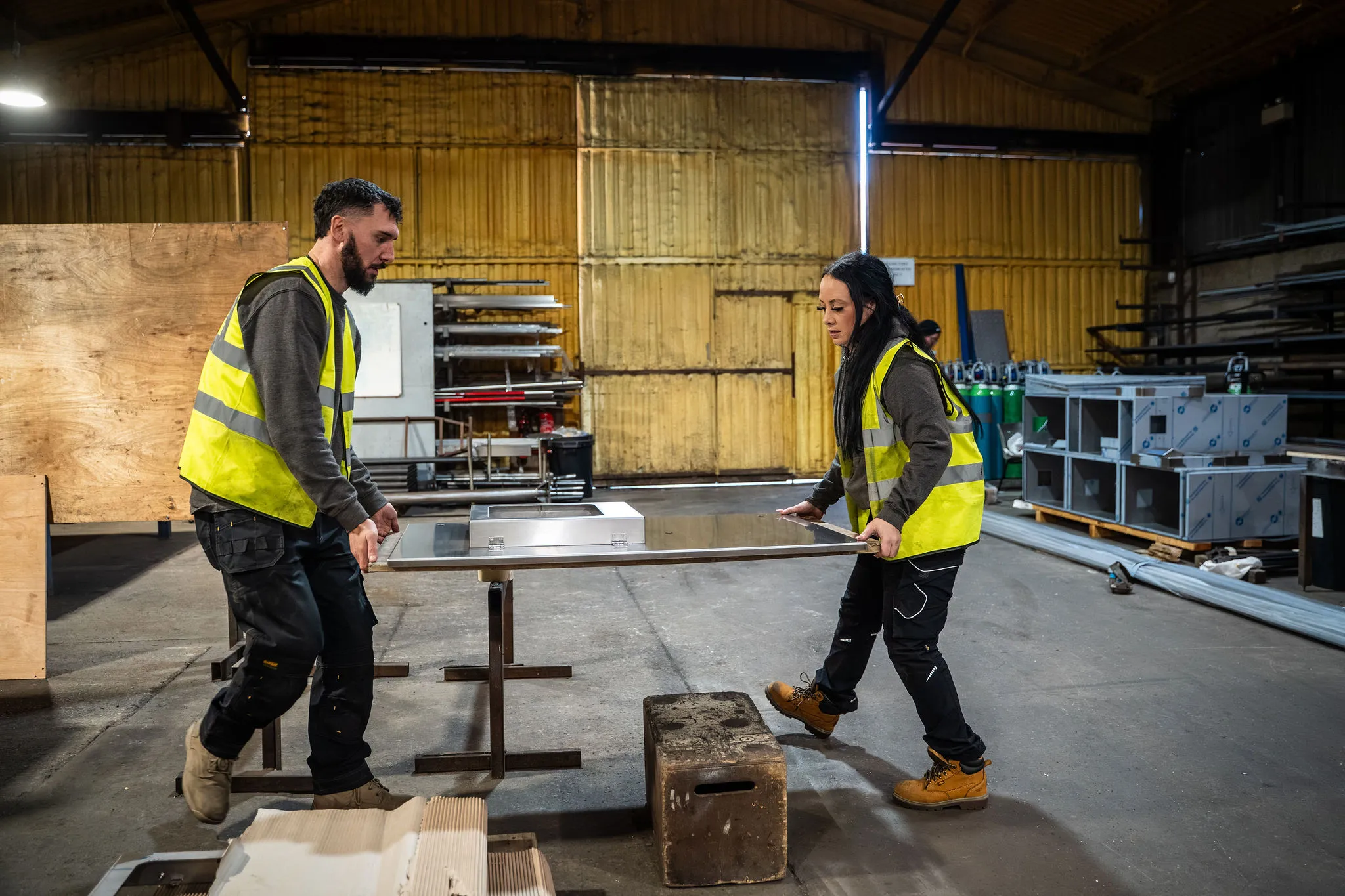 Two workers in yellow safety vests lifting a metal sheet on a stand in a workshop.