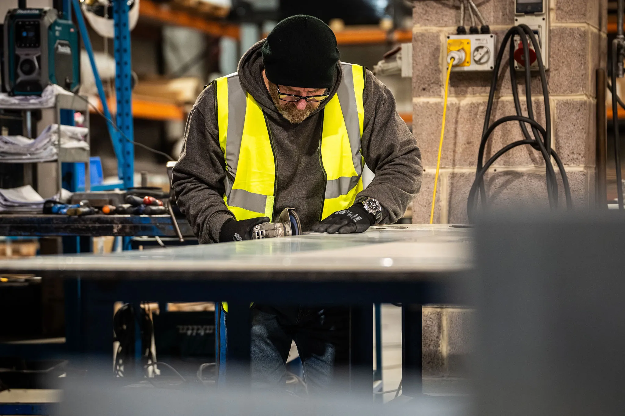 Worker wearing a yellow safety vest using a grinder on a metal sheet in a workshop.