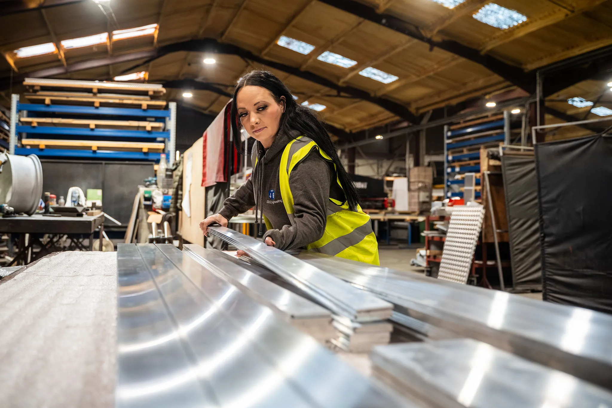 Woman in yellow safety vest handling metal sheets in a spacious industrial workshop.