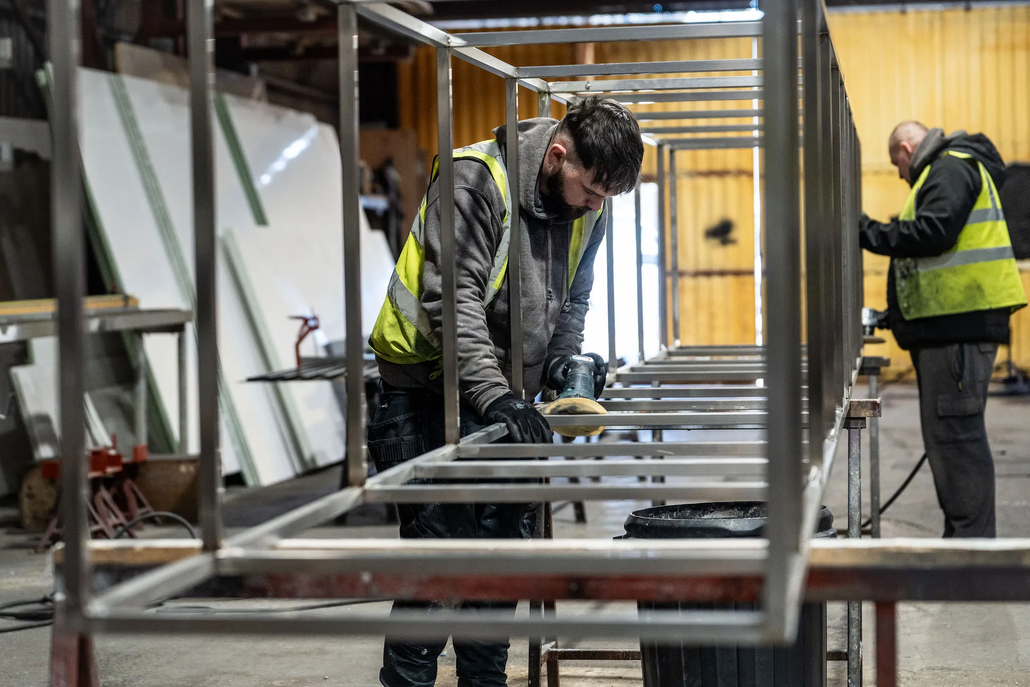 Two workers in high-visibility vests smoothing metal frame inside an industrial workshop.