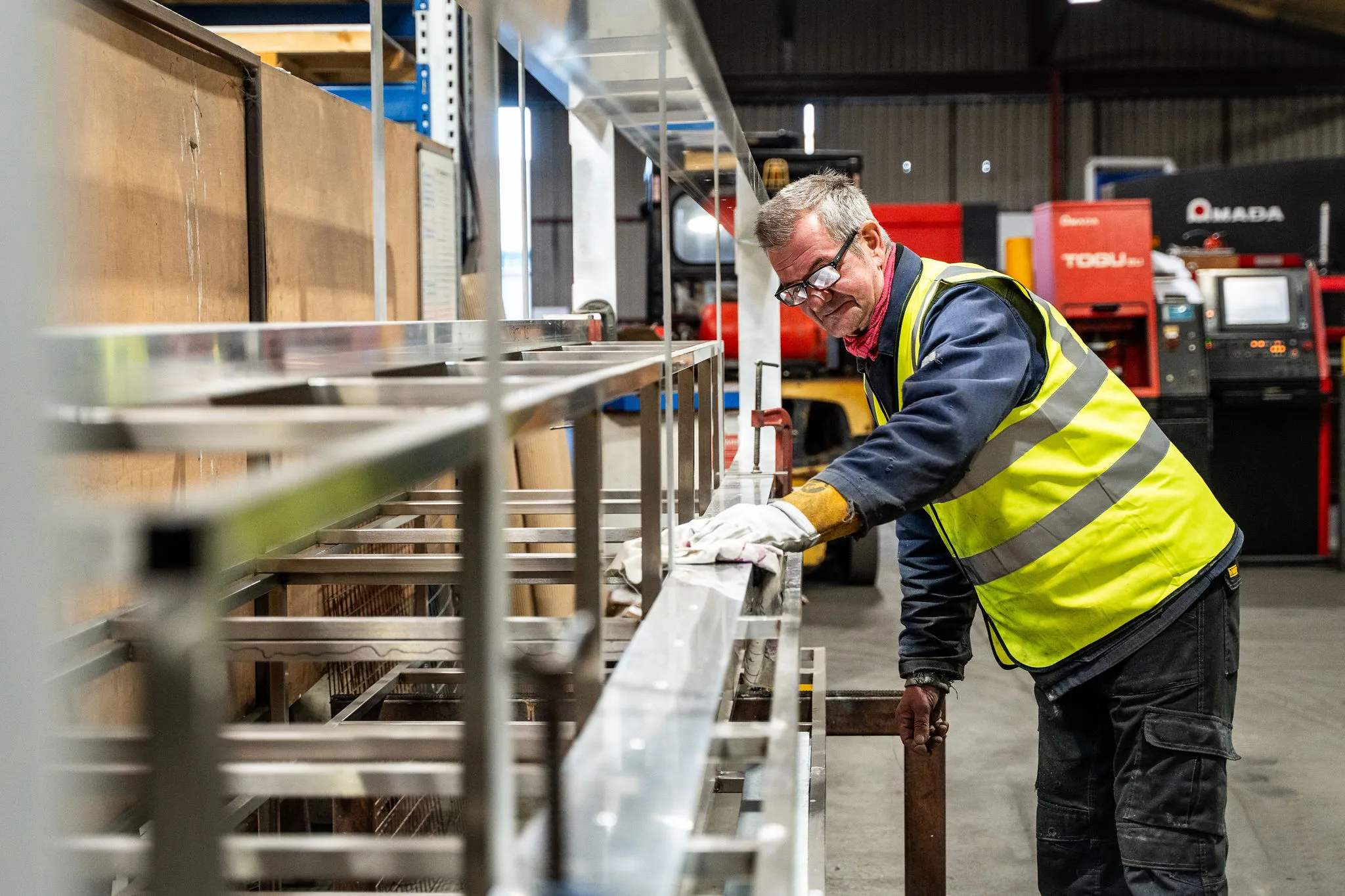 Man in a yellow safety vest working on a large metal structure inside an industrial workshop.